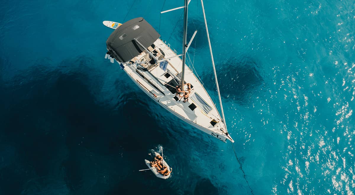 A monohull lies moored in a turquoise bay, while a few people take a swim in the water.